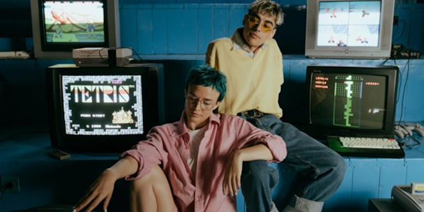 Two young people sit surrounded by vintage gaming consoles