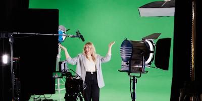A woman standing in front of a green screen, with a digital camera in the foreground