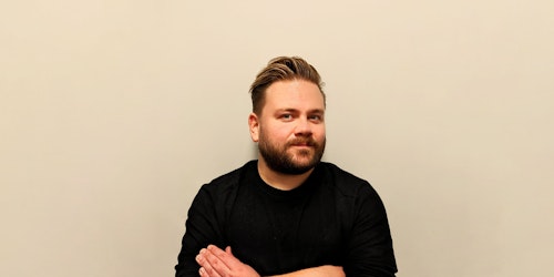A man in a black jumper standing against a beige office wall. He has styled brown hair and a beard.