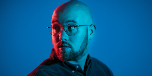 A man with a shaved head and beard wearing a dark shirt looks over his shoulder, lit by red light against a blue background.
