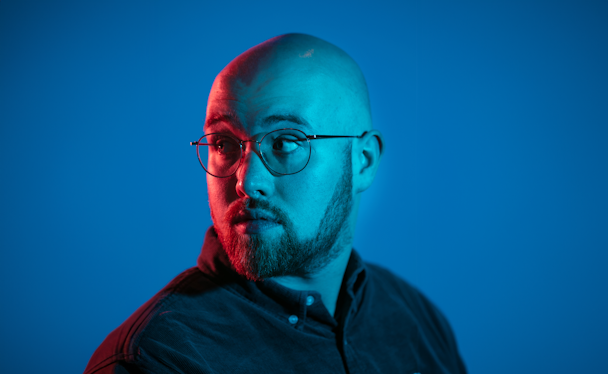 A man with a shaved head and beard wearing a dark shirt looks over his shoulder, lit by red light against a blue background.