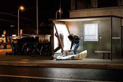 Two people wearing masks dismantle a bus stop advertising panel in the dark