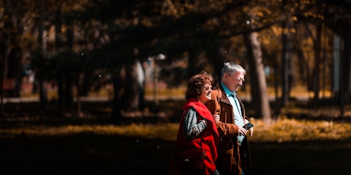 Two older people - a man and a woman - walking through a forest, with a visible cellphone in the man's hand