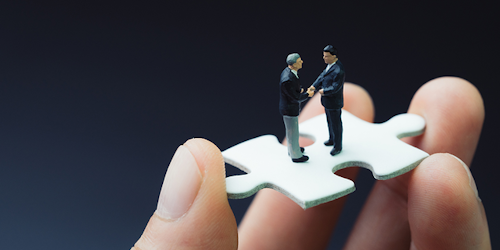 Two men shaking hands on a puzzle piece being held by a bigger hand