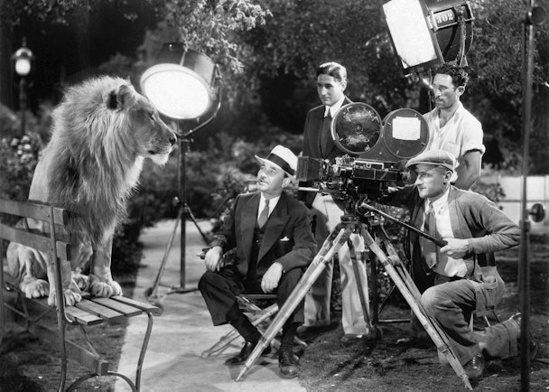 In this black and white photo, a 1930s film crew film a lion sitting on a park bench.