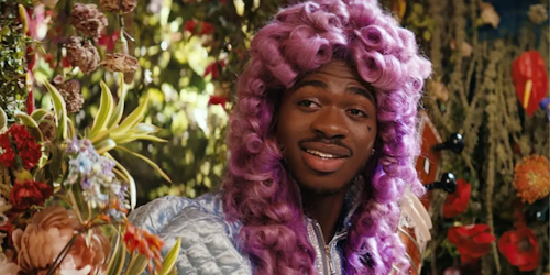 Lil Nas X wearing curly pink wig against a backdrop of flowers