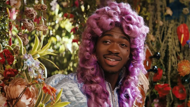 Lil Nas X wearing curly pink wig against a backdrop of flowers