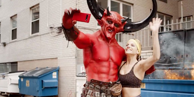 Satan and blonde woman snapping selfie in front of dumpster
