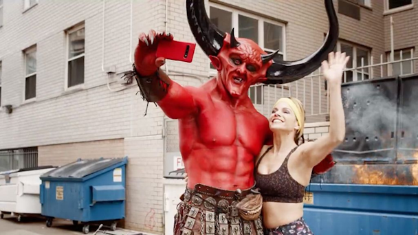 Satan and blonde woman snapping selfie in front of dumpster