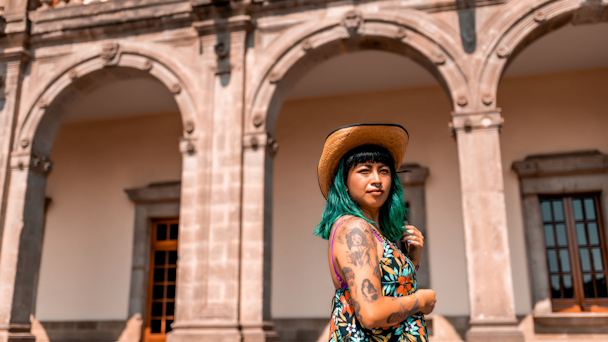 Person with tattoos and green hair standing in front of arches  