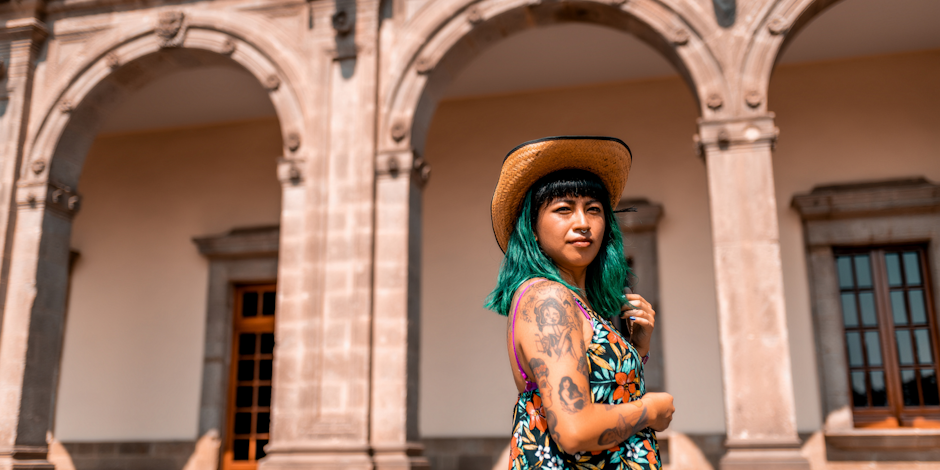 Person with tattoos and green hair standing in front of arches  
