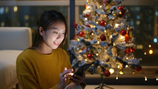 Woman on phone with white lit Christmas tree behind her Woman on phone with white lit Christmas tree behind her