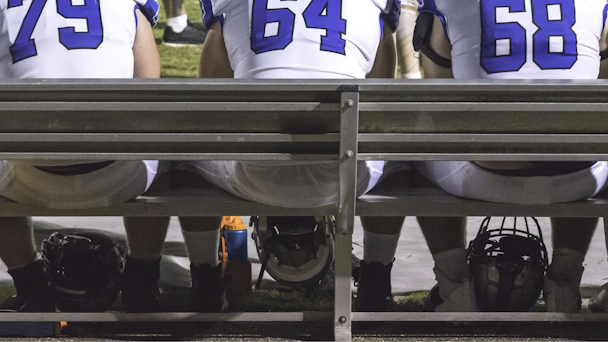 Three American football players sitting on sidelines bench with helmets between their feet