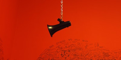 A loudspeaker dangling from a chain in front of a red wall with graffiti on it