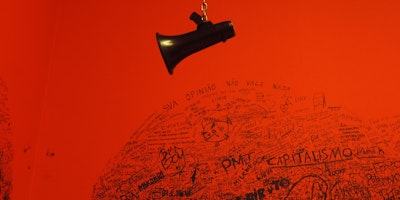 A loudspeaker dangling from a chain in front of a red wall with graffiti on it