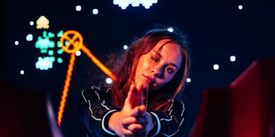 A woman with her fingers in the shape of a gun infront of gaming-style neon signs
