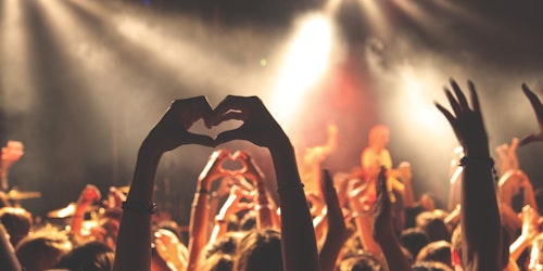 Fans at a concert; one of them holds up their hands in a love-heart shape