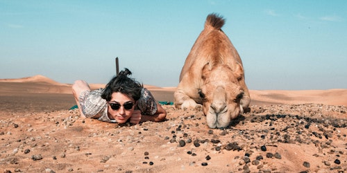 A person lying down, facing the camera - beside a camel doing the same
