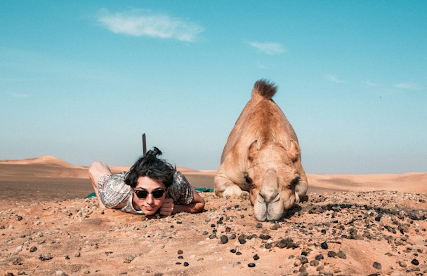 A person lying down, facing the camera - beside a camel doing the same