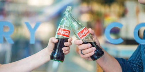 Two people, toasting with glass bottles of Coke
