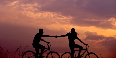 Two people, in silhouette, riding bicycles and reaching out to one another