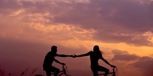 Two people, in silhouette, riding bicycles and reaching out to one another