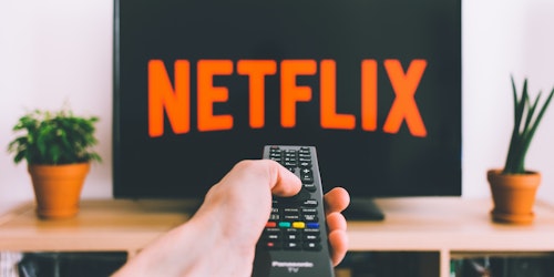 A hand holding a remote control in front of a television, turning on Netflix