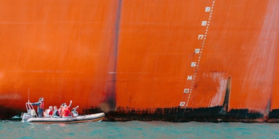 A small dinghy filled with people, dwarfed at sea by an enormous tanker