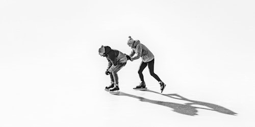 Two people in snowclothes and ice skates; one is pushing the other forward