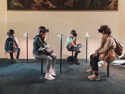 Four people wearing virtual reality headsets while sitting down on stools. A large renaissance painting hangs above them.