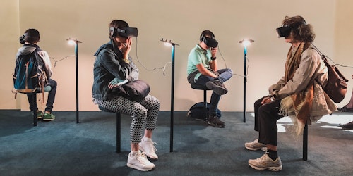 Four people wearing virtual reality headsets while sitting down on stools. A large renaissance painting hangs above them.
