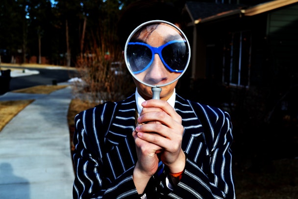 A person wearing blue spectacles, holding up a magnifying glass to their face, enlarging their features