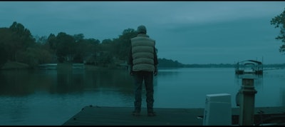 A man standing on a jetty, looking out over a still lake, with a Fincher-esque composition