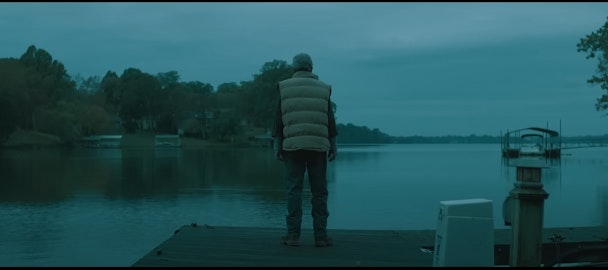 A man standing on a jetty, looking out over a still lake, with a Fincher-esque composition