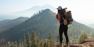 A woman atop a mountain in hiking gear, surrounded by pine trees