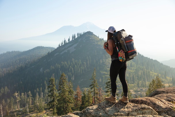 A woman atop a mountain in hiking gear, surrounded by pine trees