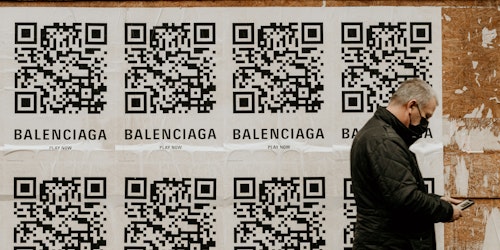 A man walking past a wall plastered with posters for Balenciaga, featuring QR codes