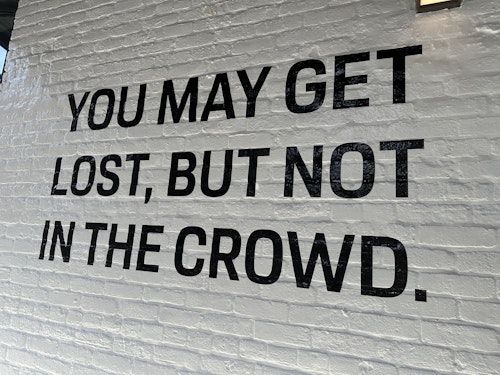 A message painted in black on a white brick wall reads 'you may get lost but not in the crowd'
