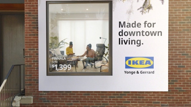 Looking through the window of an apartment building, a mother and her daughter visable inside sat on Ikea furniture, while surrounding the window outside is an Ikea billboard with the tagline 'Made for downtown living'
