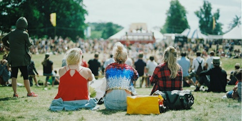 Three people sit on the grass at a festival with a big top tent in the background