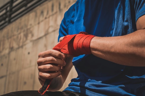 A man in a blue t-shirt wraps red tape around his knuckles as if preparing to box