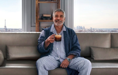 George Clooney sits on a sofa holding a cup of black coffee