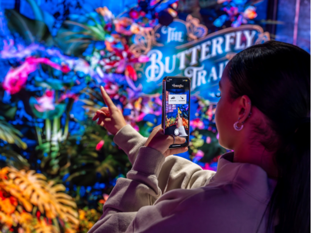 A woman with brown hair stands in front of a digital screen using her mobile phone to play an augmented reality game