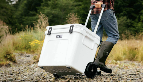 A Yeti cooler being wheeled over rough terrain by a person wearing rubber boots